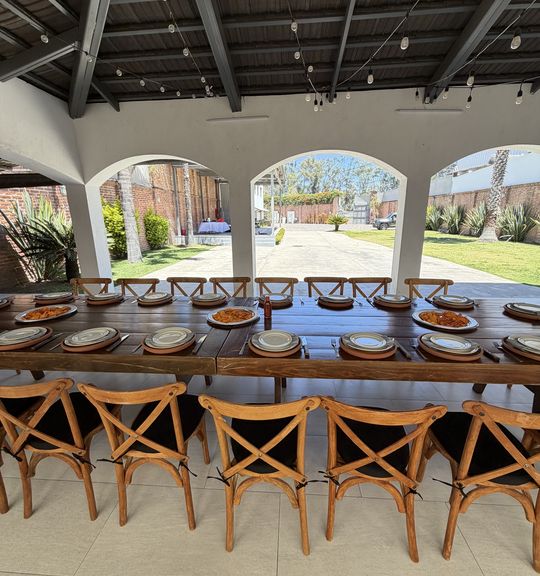 Mesa larga de madera con vajilla puesta y sillas cross-back bajo una pérgola