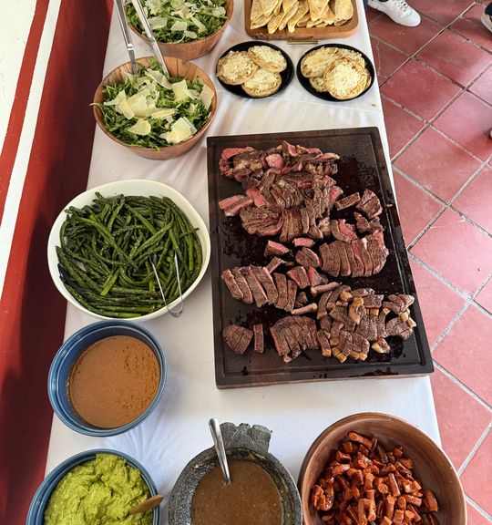 Vista cenital de la mesa montada: tabla de cortes rebanados, espárragos, ensaladas, guacamole, salsa en molcajete y chistorra